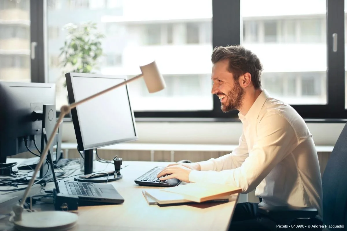 Mann im weißen Hemd sitzt vor einem Computer und lächelt - In diesem umfangreichen Beitrag erfahren Sie wissenswerte Informationen darüber, warum Büro Outsourcing immer beliebter wird.